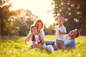 A family of four enjoys a sunny day in a grassy park, with the mother and father lying down while the children play with bubbles, the father smiling and holding a bubble wand, and the mother holding a bubble wand and a flower, surrounded by trees and a bright sun.