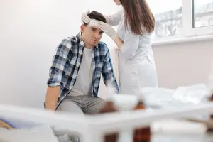 A man with a bandage on his head is sitting on a chair while a woman in a lab coat and gloves is standing behind him and probably checking his head injury.