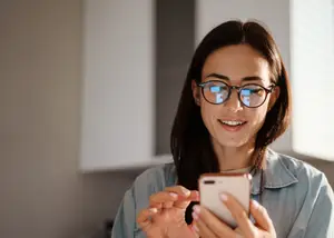 A woman with glasses is smiling and looking at her phone in a room