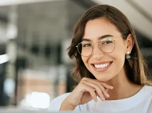 A woman with eyeglasses smiling while sitting in a chair inside an office with a blurry background
