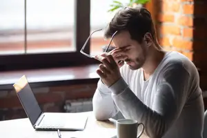 Man rubbing his eyes while sitting at a desk with a laptop and coffee cup