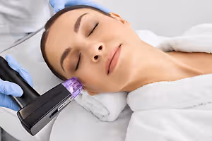 A woman is lying on a bed with her eyes closed while a person in a blue glove holds a purple-colored machine in front of her face.