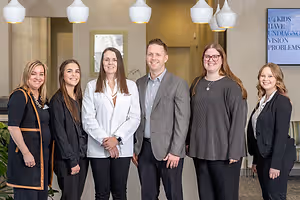 A group of six people, including a doctor, are standing together and smiling for a photo in a clinic with a digital board in the background