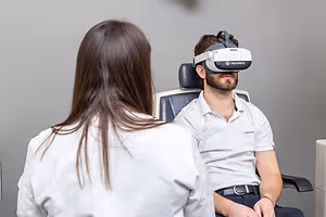 A man is sitting on a chair with a virtual reality headset, and a woman is standing behind him in a room. The man is wearing a white polo shirt and a black belt, and the woman is wearing a white coat. The man is looking at the woman, and the woman is looking at the man. Behind them is a gray wall, and a white object is on the right side of the wall.