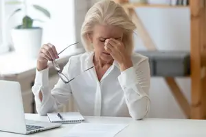 an elderly woman sitting at a desk rubbing her eyes and holding a pair of glasses