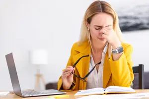 A woman sitting at a desk with a laptop and book, holding her face and wearing a yellow jacket