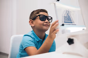 A boy wearing sunglasses is sitting on a chair and smiling while looking at a monitor and seems to be using a microscope