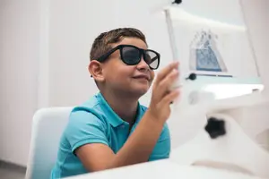 A boy wearing sunglasses is sitting on a chair and smiling while looking at a monitor and seems to be using a microscope