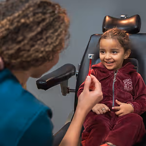 A smiling child sits in a dental chair while a dental professional brushes their teeth