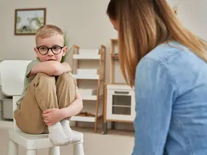 A young boy with glasses sits on a chair in a room with a woman looking at him.