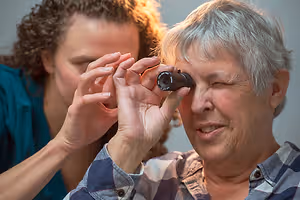 An older woman wearing glasses has her eyes examined by a young woman wearing a blue shirt.