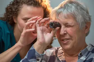 An older woman wearing glasses has her eyes examined by a young woman wearing a blue shirt.