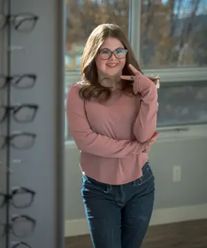 A woman in glasses is smiling and posing for a photo with her arms crossed in front of a window with a view of trees outside.