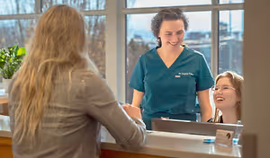 Three women stand at a reception desk. One woman in a teal uniform smiles while the other two women are also smiling.