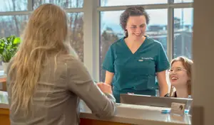 Three women stand at a reception desk. One woman in a teal uniform smiles while the other two women are also smiling.