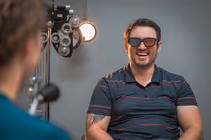 A man wearing glasses is smiling while looking at a person standing in front of an eye examination machine.