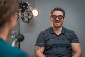 A man wearing glasses is smiling while looking at a person standing in front of an eye examination machine.