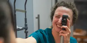 A female optometrist smiles while examining a patient's eye with a handheld ophthalmoscope