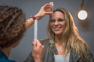 A woman is having her hair measured with a ruler by a person wearing glasses and a watch