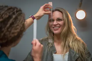 A woman is having her hair measured with a ruler by a person wearing glasses and a watch