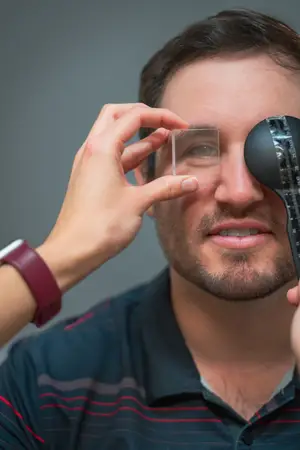 A man wearing a striped shirt is holding a clear glass and a black object with a ruler, possibly adjusting a contact lens in his eye.