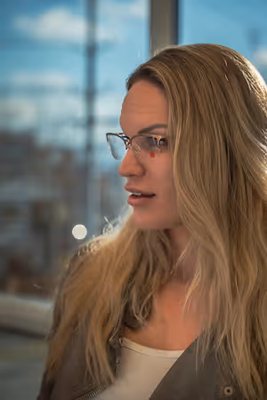 A woman with blonde hair wearing glasses and a brown jacket standing in front of a glass window.