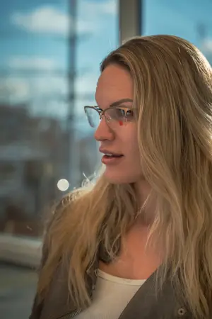 A woman with blonde hair wearing glasses and a brown jacket standing in front of a glass window.