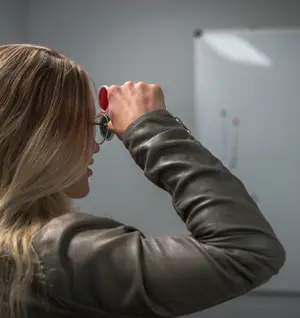 A woman with blonde hair wearing a black leather jacket and holding a pair of glasses up to her face and looking at something