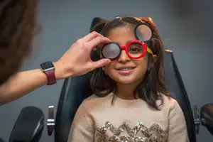 A young girl is smiling while having her eyes checked by a woman wearing a watch and bracelet