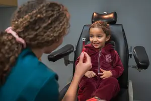 A young girl is sitting in a dental chair while a woman in a blue shirt is examining her teeth with a dental mirror