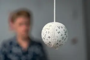A white baseball with letters and numbers is hanging from a white string with a blurred background of a person and gray wall