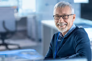 An older man wearing glasses, a blue ID, and a blue sweater is smiling while sitting on a chair in an office with a computer and a desk behind him.
