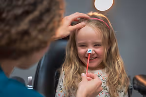 A young girl smiles and holds a toothbrush in her mouth as a woman adjusts her hair behind her head, sitting in a dental chair with a light overhead.