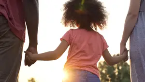 An adult man and woman holding hands with a small child standing between them while the sun shines in the background.