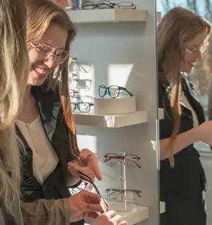 Three women wearing glasses are smiling while looking at eyeglasses in a store display