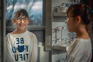 A girl in glasses stands next to another girl inside a shop with shelves of glasses behind them