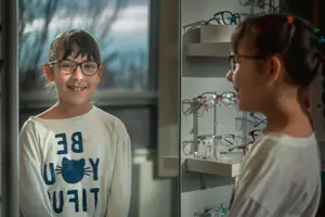 A girl in glasses stands next to another girl inside a shop with shelves of glasses behind them
