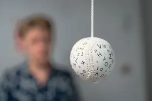 A white baseball with alphabets hanging from a white thread in front of a blurry person.