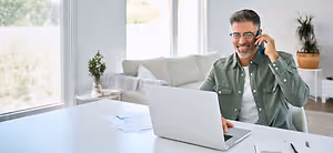 A man is sitting in a chair in front of a desk and smiling while holding a cell phone and talking to someone, and there is a laptop and papers on the desk, a couch with pillows and a potted plant on a table behind him, and a glass window with a view of trees outside
