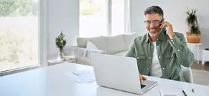 A man is sitting in a chair in front of a desk and smiling while holding a cell phone and talking to someone, and there is a laptop and papers on the desk, a couch with pillows and a potted plant on a table behind him, and a glass window with a view of trees outside