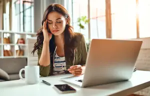 A woman is sitting on a chair in front of a desk with a laptop, cup, and pen. She is closing her eyes and putting her hand on her forehead, probably feeling stressed. She is holding her glasses in her other hand. Behind her is a shelf with some books and a couch with a pillow. There is a plant on the side of the desk.