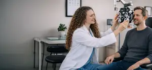 A woman is examining a man's eyes in a medical setting using an ophthalmoscope