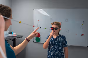 A woman and a boy are standing in front of a whiteboard with an eye chart. The woman is holding a stick with a red light, and the boy is wearing sunglasses. They are both looking at the whiteboard. The woman is pointing at the chart with her left hand, while the boy is holding the stick with his right hand. A desk with a pen and a storage box is on the left side.