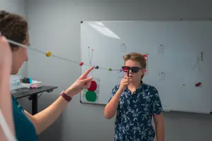 A woman and a boy are standing in front of a whiteboard with an eye chart. The woman is holding a stick with a red light, and the boy is wearing sunglasses. They are both looking at the whiteboard. The woman is pointing at the chart with her left hand, while the boy is holding the stick with his right hand. A desk with a pen and a storage box is on the left side.