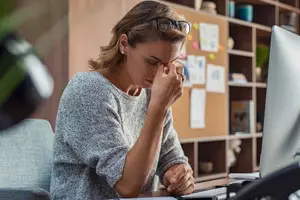 A woman is sitting at her desk, covering her eyes with her left hand, while looking down at her laptop. The image captures a moment of distress or discomfort, possibly due to eye strain or a headache. The woman is wearing a gray sweater and earrings, and her desk is cluttered with various items, including a potted plant, a cup, and a telephone. Behind her, there is a wooden shelf with several papers attached to it, and a blurry view of another person's head on the left side.