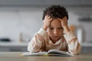 A boy sitting at a table reading a book with his hands covering his face