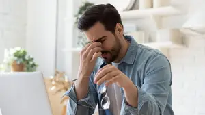 A man in a denim shirt sits in front of a laptop, holding his face in pain.