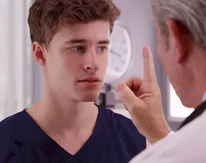 A male doctor points to a teenage boy's ear as he examines it.