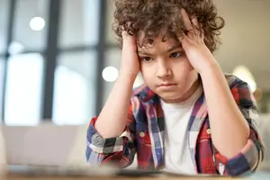 A young boy with curly hair is sitting in a room and seems to be feeling stressed. He has his hands on his head and is looking down, probably thinking about something. He is wearing a white shirt with a red and blue plaid long-sleeve shirt. Behind him is a laptop and a glass window with a view of the outside. There is a bright light on the right side of the room.