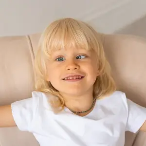 A young girl with blonde hair and blue eyes is lying on a beige couch and smiling at the camera. She is wearing a white t-shirt and a necklace.